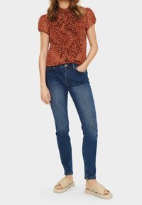 Rostfärgad blus med volangdesign och vita prickar, i kombination med figurnära mörkblå jeans och beige sandaler med strukturerade remmar.