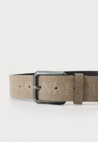 Beige mockaskärp med en mattsilver rektangulär spänne. Har flera hål för justering och ett varumärkesdetalj nära spännet.