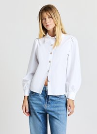 Camicia bianca con colletto, maniche a sbuffo e dettagli in bottoni argentati, abbinata a jeans di denim azzurri. Tessuto dalla consistenza liscia.