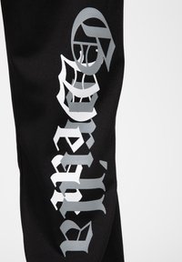 Black fabric pant featuring large silver graphic text and motifs. Text is stylized, with a textured finish, on a breathable material.