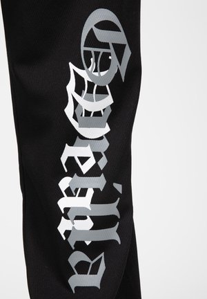 Black fabric pant featuring large silver graphic text and motifs. Text is stylized, with a textured finish, on a breathable material.