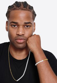 Gelaagde sieraden set met een lange zilveren kraalhalsketting, een witte armband met een gouden accent en een zwart T-shirt.