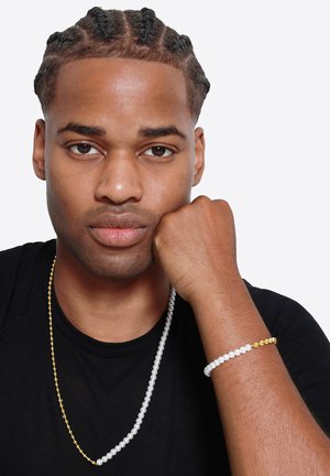 Ensemble de bijoux en perles, comprenant un long collier en perles argentées, un bracelet blanc avec un accent doré, et un t-shirt noir.