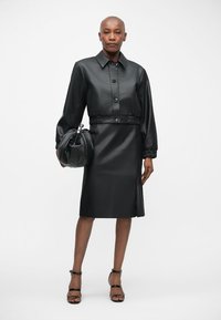 Crna kožna haljina s kragnom na dugme, elastičnom strukom i dugim rukavima. Uparena s okruglom crnom torbom i sandalama s remenčićima.