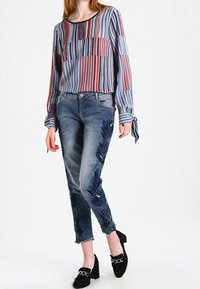 Maglia a strisce blu e rossa con maniche lunghe e polsini arricciati, abbinata a jeans blu sdruciti con ricami di foglie verdi sui lati. Scarpe nere con tacco.