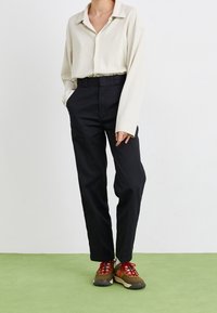 Chemise beige à manches longues avec patte de boutonnage, associée à un pantalon noir cintré et des baskets marron avec des lacets rouges et des semelles texturées.