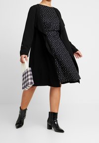 Schwarzer offener Mantel über einem navyblauen Kleid mit Punkten. Model trägt eine kleine, strukturierte Tasche mit lila und weißen Perlen. Schwarze Ankle Boots.