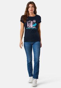 T-paita, jossa on tummansininen puuvilla, etupuolella kuvio, jossa näkyy vuoria, puita ja haisunäätä pinkissä ja sinisessä värissä, yhdistettynä sinisiin farkkuihin.