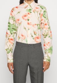 Camisa de manga larga con botones y estampado de flores en rosa y melocotón, combinada con pantalones gris ajustados, sobre una persona de pie.