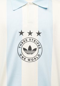 Πολό πουκάμισο με κατακόρυφες ρίγες σε ανοιχτό μπλε και λευκό, με κεντημένα τρία μαύρα αστέρια και κυκλικό λογότυπο που αναγράφει «Three Stripes One World.»