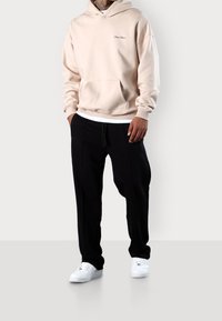 Beżowa bluza z kapturem z przednią kieszenią i białym logo, w połączeniu z czarnymi spodniami dresowymi z elastycznym paskiem, noszona z białymi trampkami.