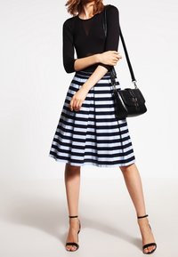 Donna che indossa un top nero a maniche lunghe, una gonna al ginocchio a righe bianche e nere, scarpe con tacco alla caviglia nere e porta una borsa nera a spalla.