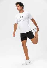 Hvit tettsittende treningsskjorte med svart logo, sammen med svarte shorts. Modellen strekker seg, iført hvite joggesko med grå detaljer.