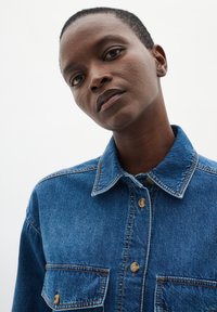 Camicia di jeans di blu medio, con colletto, due tasche sul petto e bottoni marroni. Il tessuto presenta una trama testurizzata con cuciture visibili.