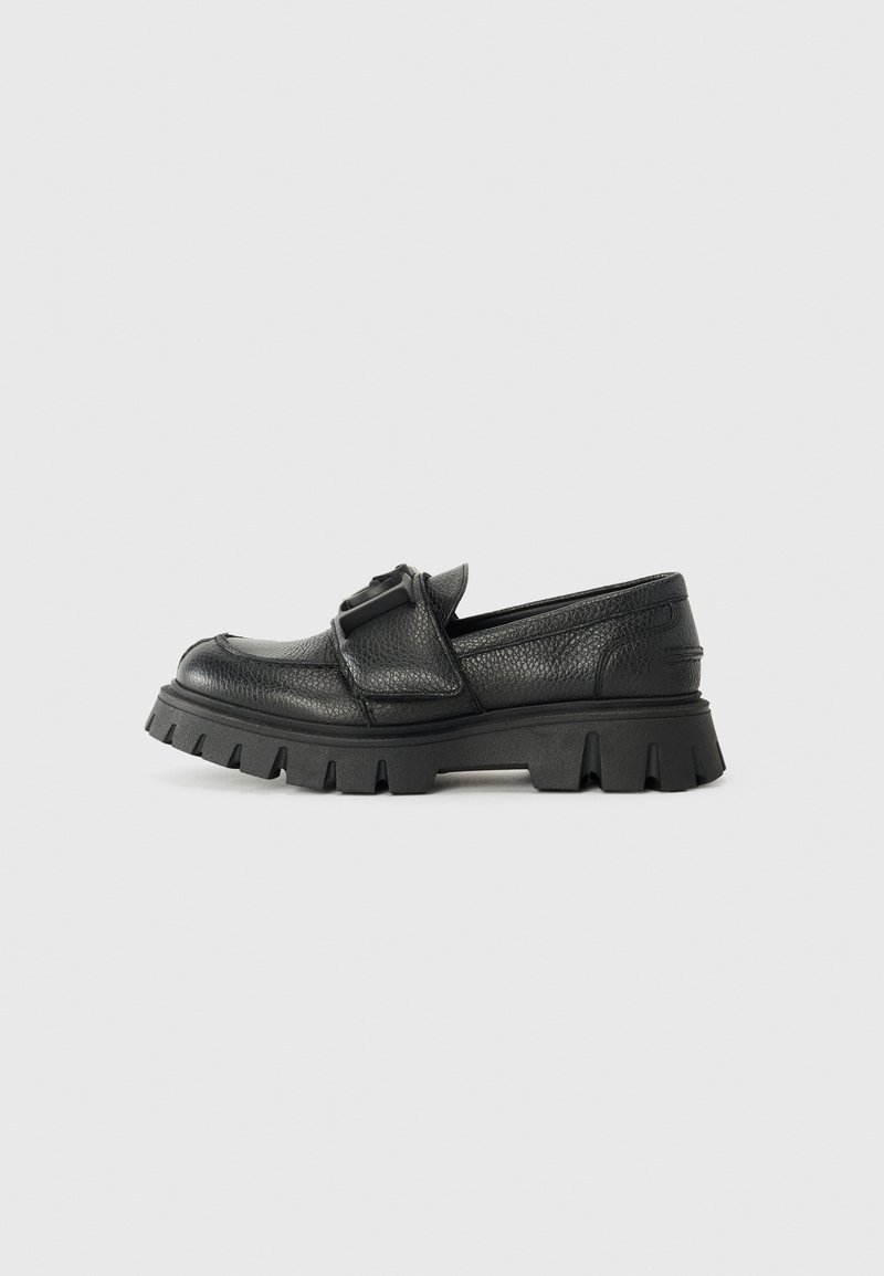 Chaussure noire en cuir à enfiler avec une surface texturée et une semelle en caoutchouc épaisse présentant de profondes rainures. Détail d'accent avec boucle ajustable.