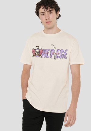 Young man with curly hair wearing cream T-shirt with colorful "One Piece" skull logo and black pants, standing against plain background.