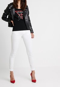 Veste de moto en cuir noir, débardeur noir décontracté avec un logo, jeans skinny blancs et escarpins rouges sur un fond neutre.