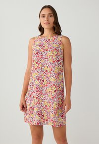 Vrouw staat rechtop en draagt een mouwloze, knielange jurk met een rood, geel en groen bloemenpatroon op een witte achtergrond.