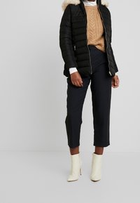 Černá péřová bunda s kapucí z umělé kožešiny, béžový svetr s copánkovým vzorem a kárované černé kalhoty, doplněné krémovými kotníkovými botami.