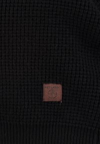 Tessuto di maglia nera con una finitura a coste e un'etichetta in pelle marrone con la scritta "KEY LARGO" che presenta un design del logo.