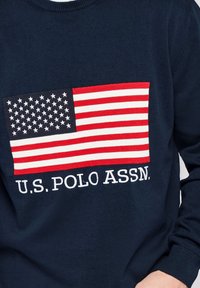 Pull bleu marine avec un grand écusson du drapeau américain et un texte blanc indiquant "U.S. POLO ASSN." sur la poitrine.
