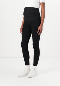 Black maternity leggings with a stretchy, over-the-bump waistband. Smooth texture, fitted design, and ankle-length cut, paired with white sneakers.