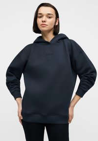 Νεαρή γυναίκα με κοντά σκούρα μαλλιά, που φοράει μια φαρδιά σκούρα μπλε μπλούζα με κουκούλα, με τα χέρια στη μέση, σε απλό φόντο.