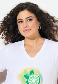 Camiseta de algodón blanca con cuello en V, que presenta un colorido estampado de cítricos con hojas verdes y texto. El modelo lleva pendientes de aro y tiene el cabello rizado.