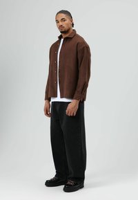 Bruine overhemd met knopen en lange mouwen over een wit T-shirt, gecombineerd met wijde zwarte broek en zwarte schoenen met roze veters.