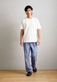 Λευκό κοντομάνικο μπλουζάκι, μπλε ριγέ παντελόνι και σκούρα παπούτσια. Το παντελόνι διαθέτει κάθετες ρίγες και έχει λεία υφή. Ουδέτερο φόντο.