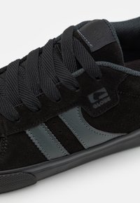Black suede skate shoe with gray leather accents, featuring rounded toe, six eyelets, and textured rubber sole. Brand logo displayed.