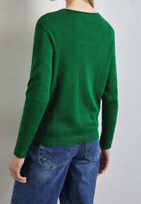 Pull en maille vert avec col rond et manches longues, doté d'un ourlet et de poignets côtelés. Porté avec un jean en denim bleu. Texture lisse.