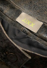Veste en cuir marron foncé usée avec doublure noire et étiquette beige cousue à l'intérieur, portant l'inscription "JJXX" en lettres jaunes.