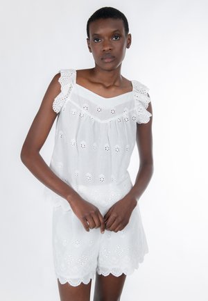 Jeune femme noire portant un haut sans manches en dentelle ajourée blanche et un short assorti, debout devant un fond blanc uni.