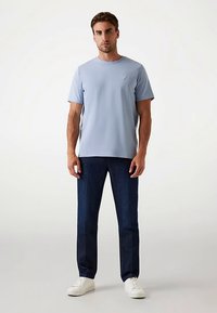 T-shirt bleu clair à manches courtes avec un col rond, associé à un jean bleu foncé et des baskets blanches, posé devant un fond neutre.