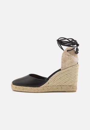 Espadrille compensée en cuir noir avec semelle en jute tissé et tissu beige texturé au talon. Comprend des liens réglables en cordon noir à la cheville.