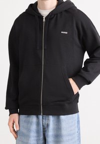 Černá mikina na zip z měkké tkaniny, s přední kapsou, šňůrkami a malým bílým logem na levé straně hrudi.