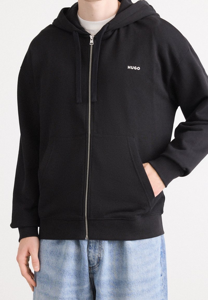Černá mikina na zip z měkké tkaniny, s přední kapsou, šňůrkami a malým bílým logem na levé straně hrudi.