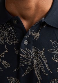 Marineblå poloshirt med lys beige blomsterprint, tekstureret stof, tre-knaps slids og korte ærmer ved kraven.