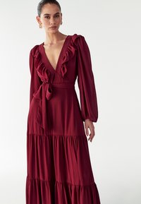 Bordo spalvota ilgos rankovės suknelė su raukinėlių apykakle, liemens raiščiu ir pakopine sijona. Medžiaga atrodo lengva ir tekstūruota.