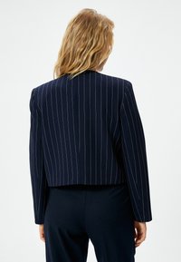 Tumman siniharmaa raidoitettu bleiseri, jossa on lyhennetty malli, istuva leikkaus ja rakenteelliset olkatuhdotteet. Kangas näyttää sileältä ja siinä on hienovarainen kiilto.