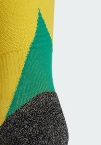 Kolorowe skarpetki mają żółty czubek, zielony trójkątny wzór oraz teksturowaną czarną stopę. Materiał wydaje się miękki i dziany.