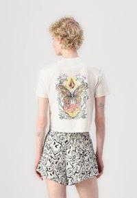 Svetlý krémový crop top s grafickým motívom motýľa a kvetov na chrbte, skombinovaný s abstraktne vzorovanými šortkami v čierno-bielej farebnej kombinácii.