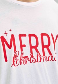 Witte katoenen T-shirt met grote rode tekst die "VROLijk kerstfeest" zegt, omringd door decoratieve sterren voor extra nadruk.