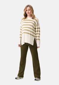 Vrouw in een crèmekleurige en olijfgroene gestreepte oversized trui, olijfgroene geribbelde broek en witte sneakers, staand tegen een eenvoudige witte achtergrond.