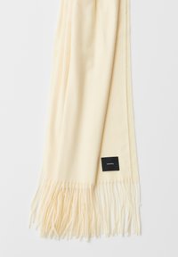 Sjaal in roomkleur, gemaakt van zacht materiaal, met franjes aan de uiteinden en een klein zwart label met witte tekst aan één rand.