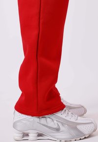 Pies en pantalones rojos que llevan zapatillas Nike Shox blancas y plateadas con suelas visibles similares a muelles sobre un fondo blanco.