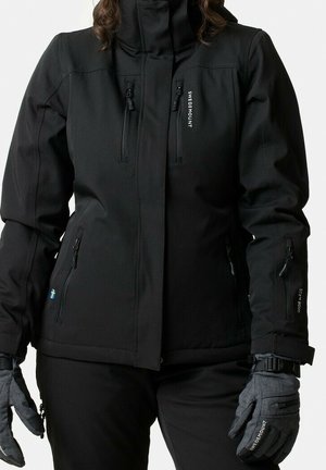 Veste d'extérieur noire avec plusieurs poches zippées, pantalon noir assorti et gants gris avec des sangles au poignet et logos de la marque.