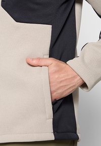 Main placée à l'intérieur de la poche avant beige et noire texturée d'une veste ou d'un pull à manches longues.