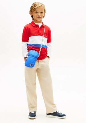 Jeune garçon portant un polo rouge, blanc et bleu, un pantalon beige, des baskets bleu marine et un petit sac bleu bandoulière, debout devant un fond blanc.